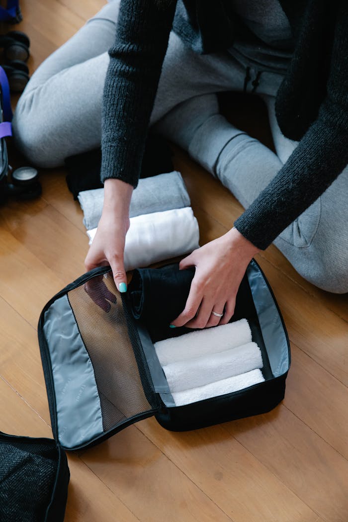 Person packing rolled towels into a pouch on wooden floor, preparing for travel.