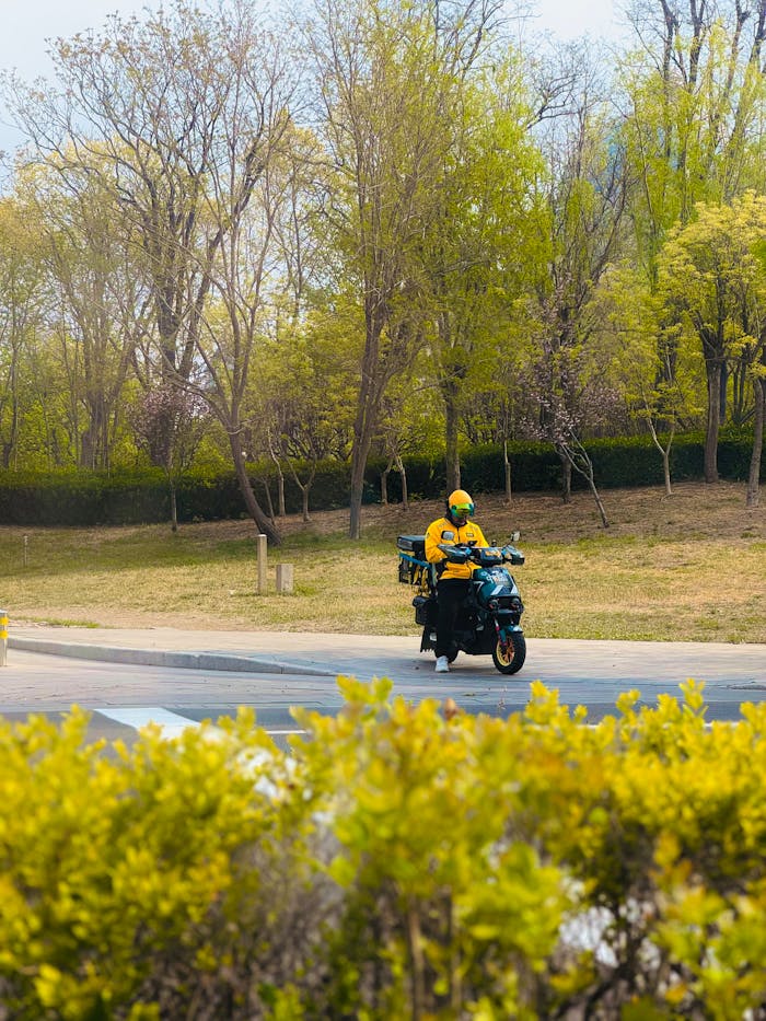 A courier in yellow uniform rides a scooter through a sunny park.