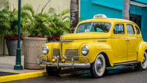 Vintage yellow taxi car parked on an urban street with lush green plants and palm trees.