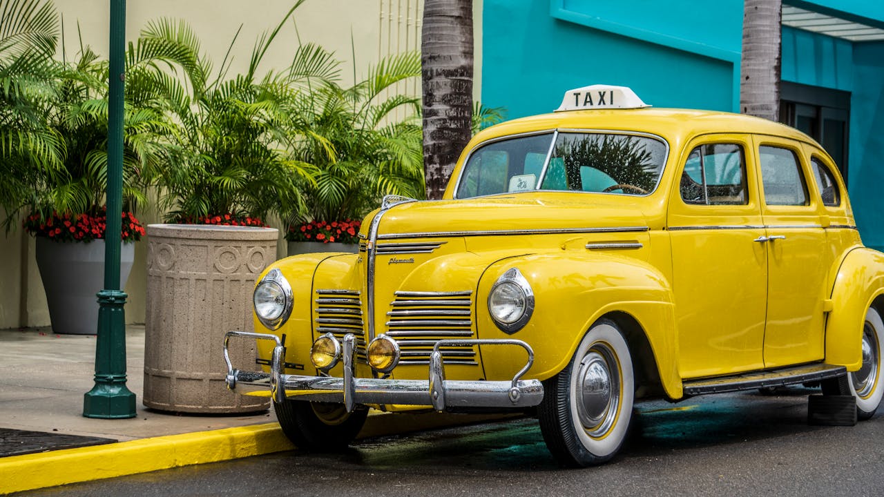 Vintage yellow taxi car parked on an urban street with lush green plants and palm trees.