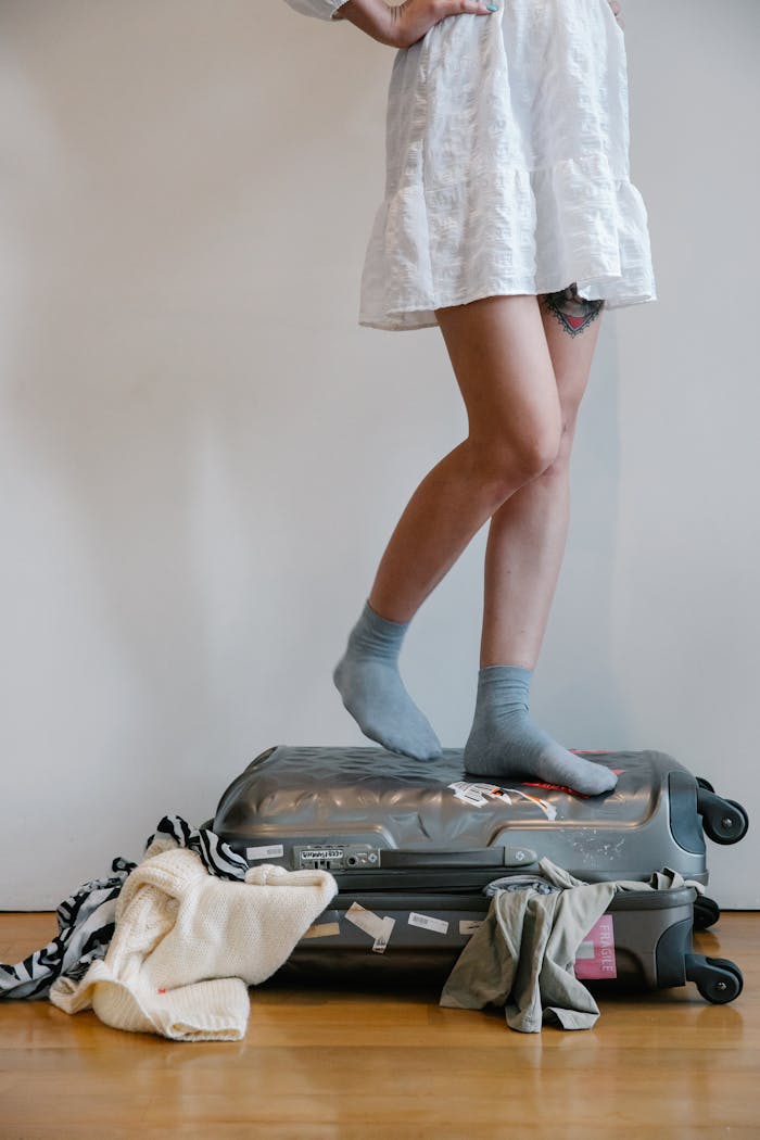 A woman attempts to close an overloaded suitcase by standing on it, surrounded by clothes.