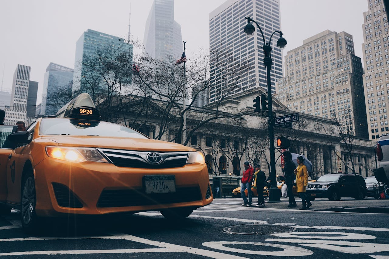 A bustling New York City street intersection featuring a yellow taxi and pedestrians with skyscrapers in the background.